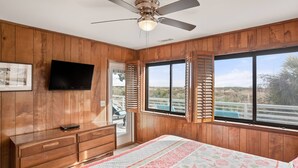 A bedroom with wooden paneling, a bed with a floral quilt, a ceiling fan, a TV mounted on the wall, and a window with shutters. - "Fant-A-Sea": Oceanfront Escape with Stunning Views, Screened Porch, Deck & Beach Access! (Saint Helena Island)