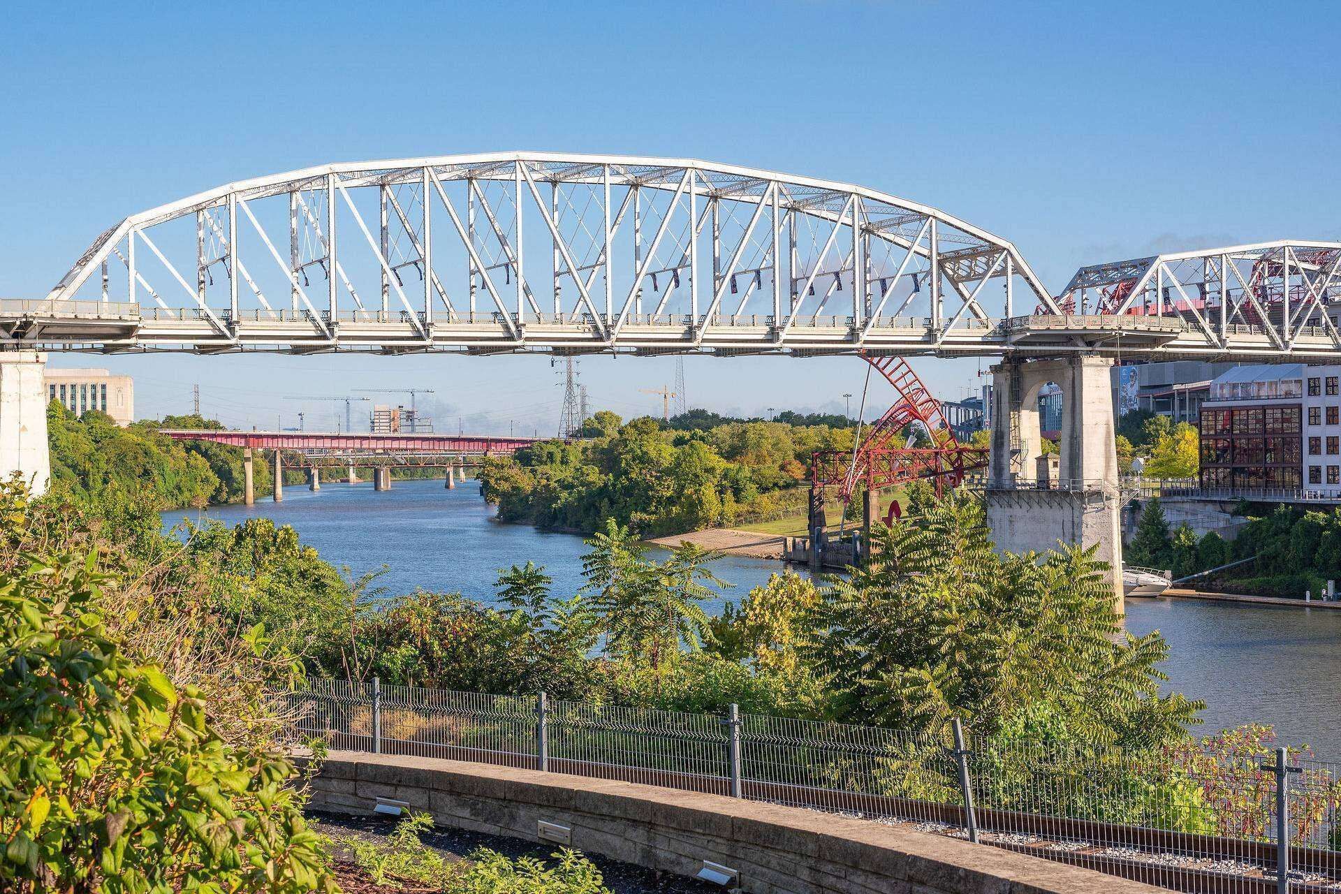 Un pont en acier doté d’une façade blanche, une rivière en contrebas et une zone délimitée par une clôture, avec de la végétation au premier plan.