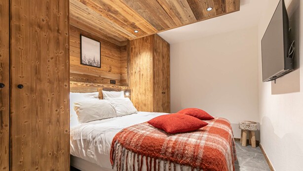 Een slaapkamer met een houten plafond, een bed met wit beddengoed en rode kussens, een klein krukje en een tv aan de muur.