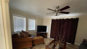A living room with a brown sofa, a glass coffee table, a television on a stand, a ceiling fan, and a window with blinds.