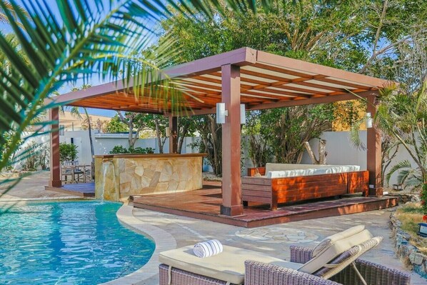Un espace aménagé au bord de la piscine, avec une pergola en bois, un banc en pierre et des chaises longues.