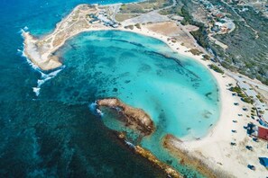 Une vue aérienne d’une zone côtière avec une plage de sable, des eaux d’un bleu limpide et quelques bâtiments dispersés.