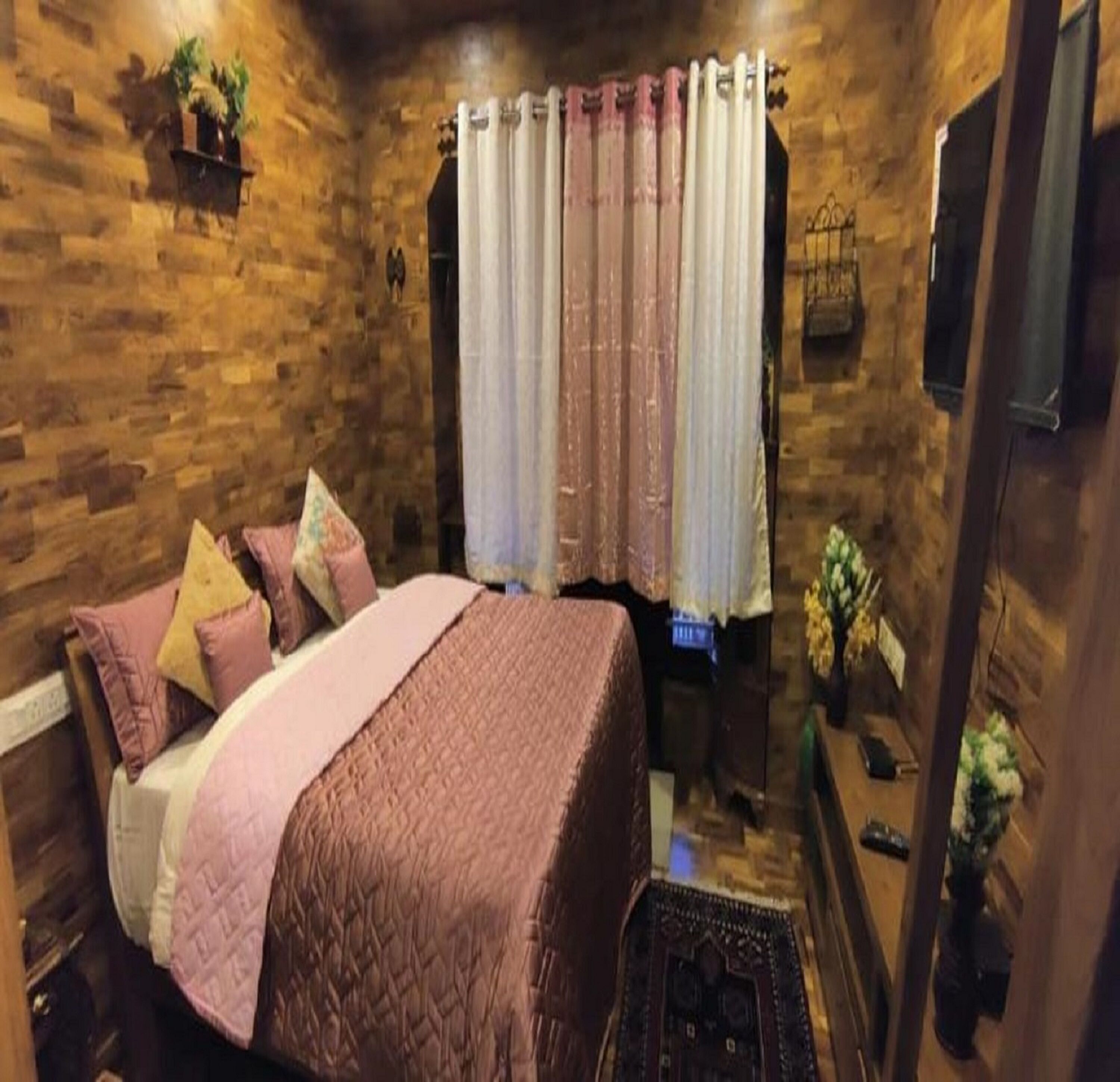 Un dormitorio con pared de madera, una cama con ropa de cama rosa, una ventana con cortinas y una mesita con un jarrón.