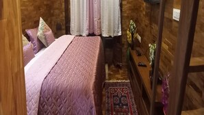 A bedroom with a wooden wall, a bed with a patterned bedspread, a nightstand with a vase of flowers, and a TV mounted on the wall. - Kasal's Stay (Kodaikanal)