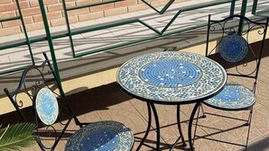 Ein runder Gartentisch mit Sitzgelegenheiten aus Mosaikfliesen, einem Metallstuhl und einem Metallzaun im Hintergrund.