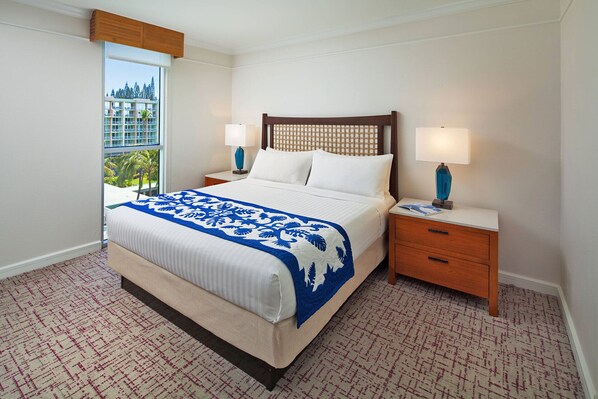 Habitación de hotel con una cama grande, dos mesitas de noche, una ventana con vistas a un edificio y palmeras, y una alfombra estampada.