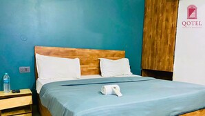 Una habitación de hotel con una cama de madera, dos almohadas blancas, una colcha azul, una botella de agua y un control remoto sobre una mesita de noche de madera.