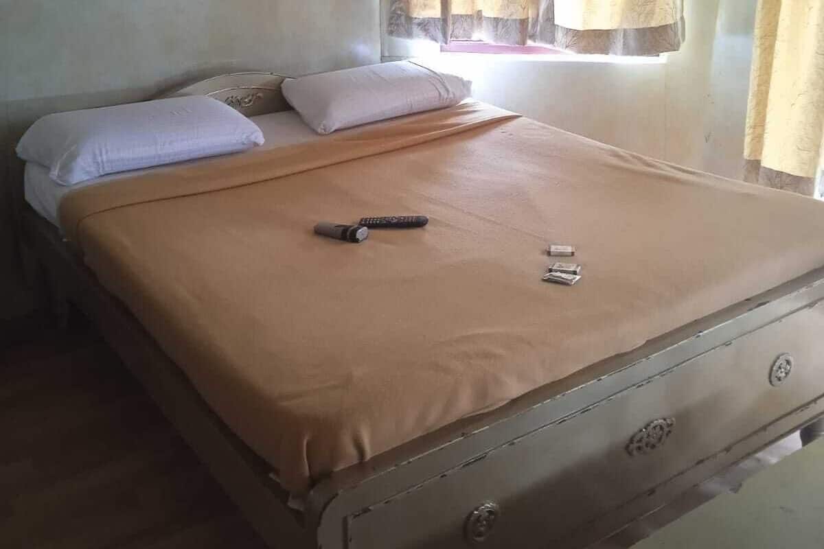 A single bed with a beige bedspread, a remote control, and a small item on the bed. There is a window with curtains, a wall with a slight discoloration, and a small table in the corner.
