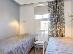 Una habitación con dos camas, una mesita pequeña, una ventana con cortinas y una lámpara de pared.