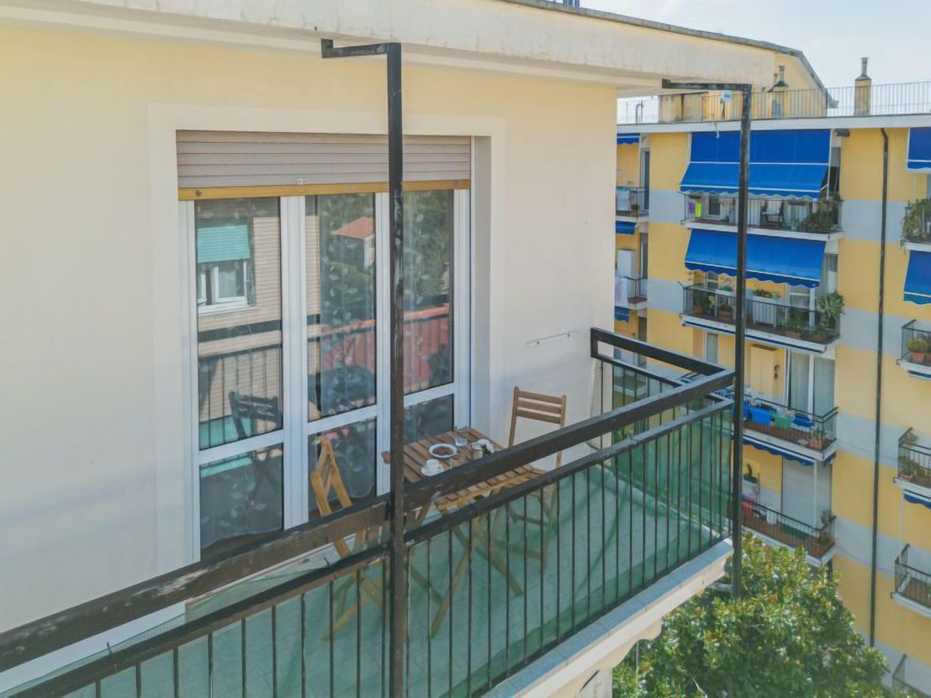 A balcony with a table and chairs, a window with a view of neighboring buildings, and a door to the building's interior.