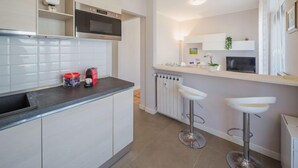 A modern kitchen with white cabinets, a black countertop, and a built-in microwave. There are two white bar stools and a white radiator. - Apartment Enrico by Interhome (Chiavari)