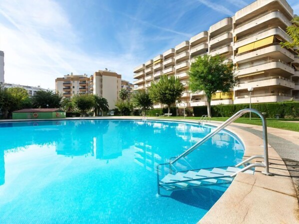 A swimming pool with a clear blue water surface, surrounded by a concrete deck and metal railings. In the background, there are multi-story buildings with balconies and a green lawn area.