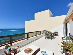 Ein Balkon mit Meerblick, ein Tisch mit Stühlen und ein kleiner Gartenbereich mit Pflanzen und einem Hut.