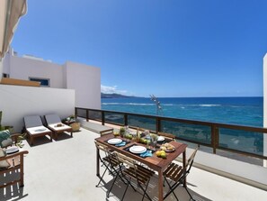 Eine Terrasse mit Esstisch für zwei Personen, mit Blick auf das klare blaue Meer und eine Windkraftanlage in der Ferne.