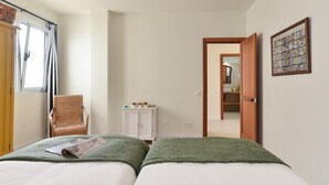 Quarto com duas camas, uma cadeira, um pequeno armário e uma porta que dá para outro cômodo.