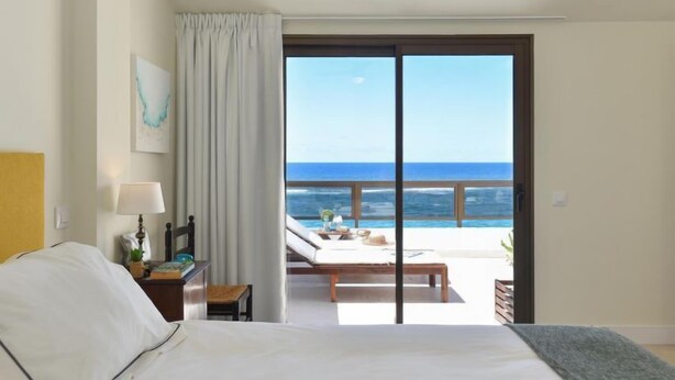 Quarto de hotel com cama, mesa de cabeceira, luminária, janela com vista para o oceano e varanda com mobiliário.
