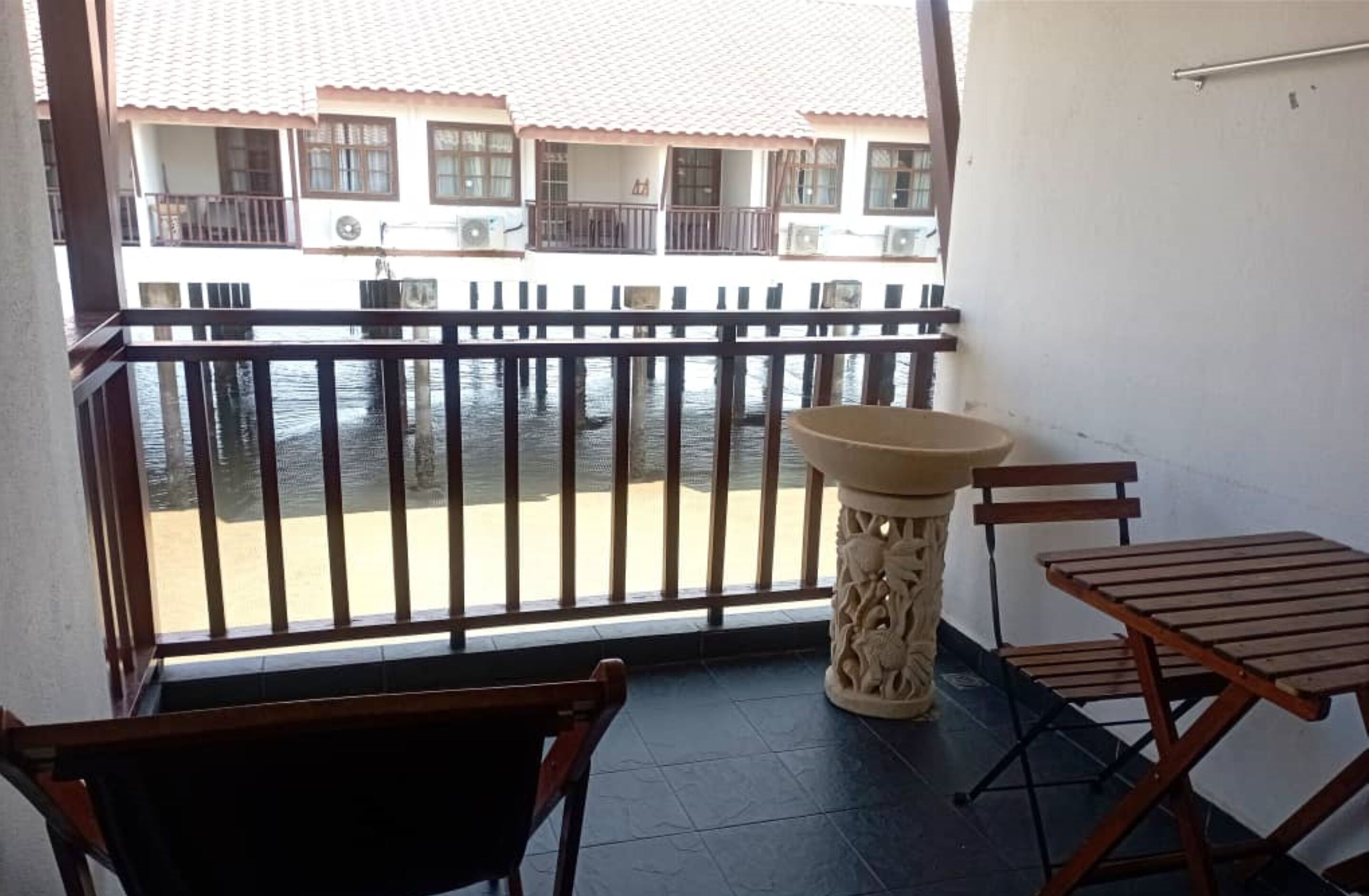A balcony with a view of a water body and buildings. There is a wooden chair, a small table, and a decorative pillar.
