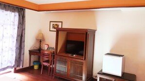 A room with a wooden TV stand, a desk with a lamp, a small refrigerator, and a window with curtains. - Alpine LXPD Premium Water Chalet (Port Dickson)