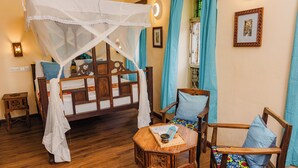 A room with a wooden bed with a canopy, two wooden chairs with cushions, a small wooden table with a book and a cup, and a wall-mounted shelf with books. - Emerald Palm Boutique Hotel (Zanzibar Town)