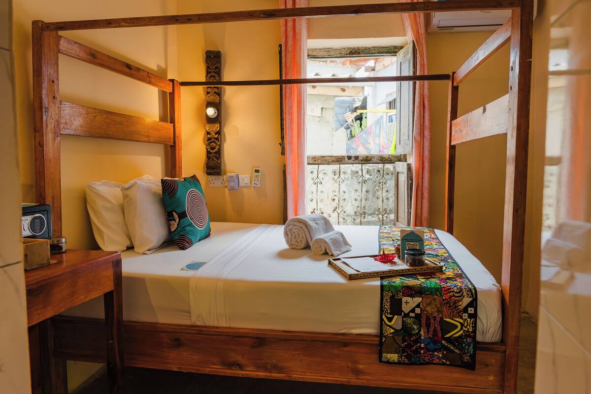 Una cama con dosel de madera, con un edredón blanco, almohadas y una toalla enrollada. Un radio y un libro están sobre la mesita de noche junto a la cama.