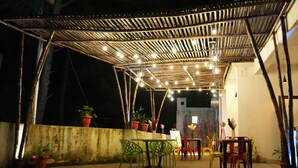 Um pátio externo com pérgola de bambu, plantas em vasos e uma mesa com cadeiras.