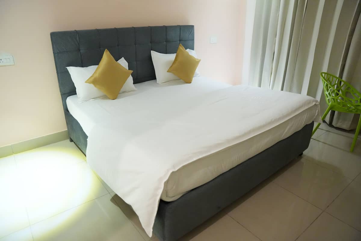 Cama bem arrumada com cabeceira escura e duas almofadas, uma cadeira verde e uma janela com persianas.