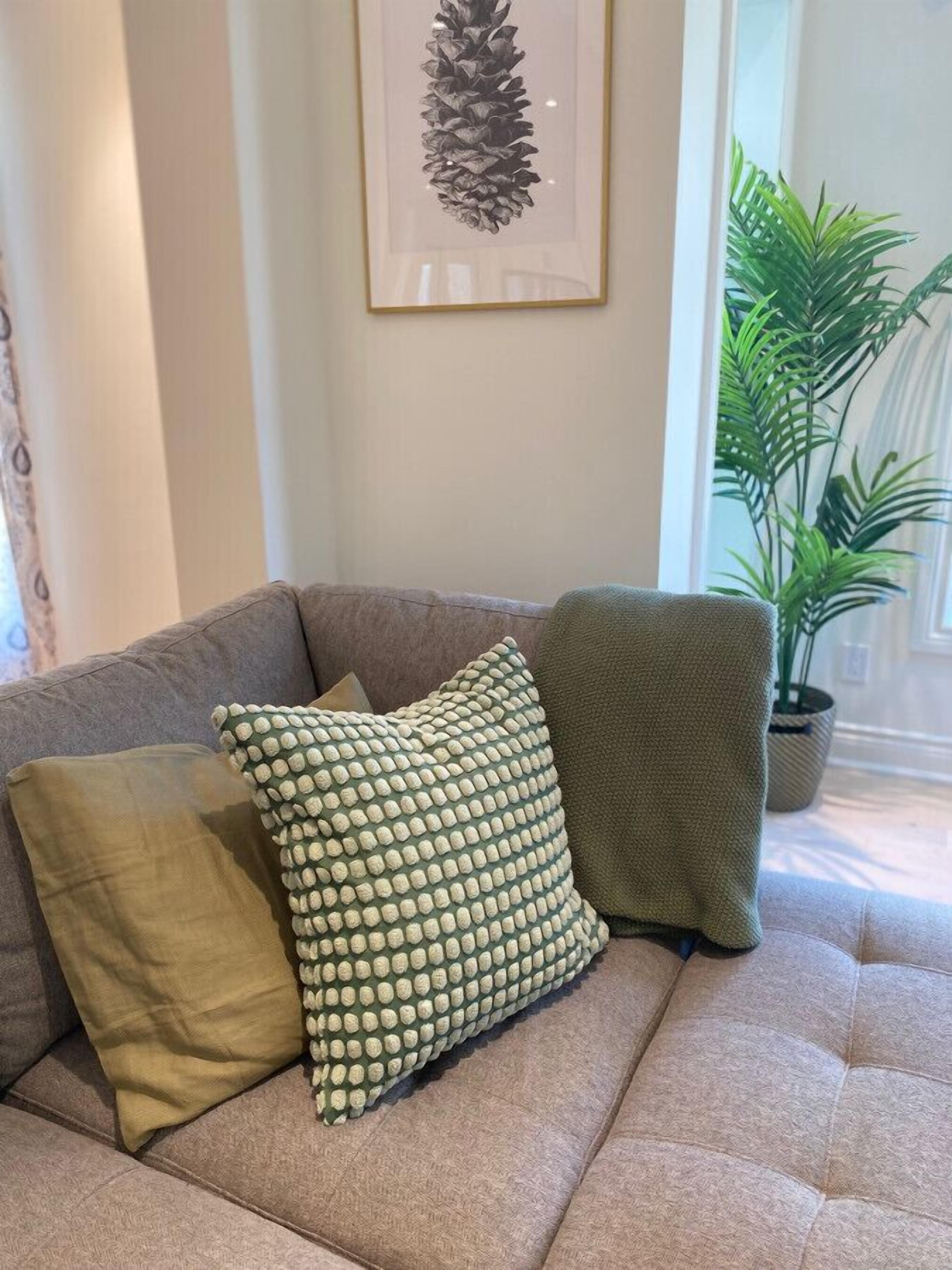 Un sofá color arena con tres almohadas, un cuadro de un pino enmarcado en la pared y una planta en maceta al fondo.