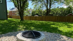 A stone fire pit with a metal screen in a backyard with a wooden fence and trees. - Downtown Picton, 3 bedroom. (Prince Edward)