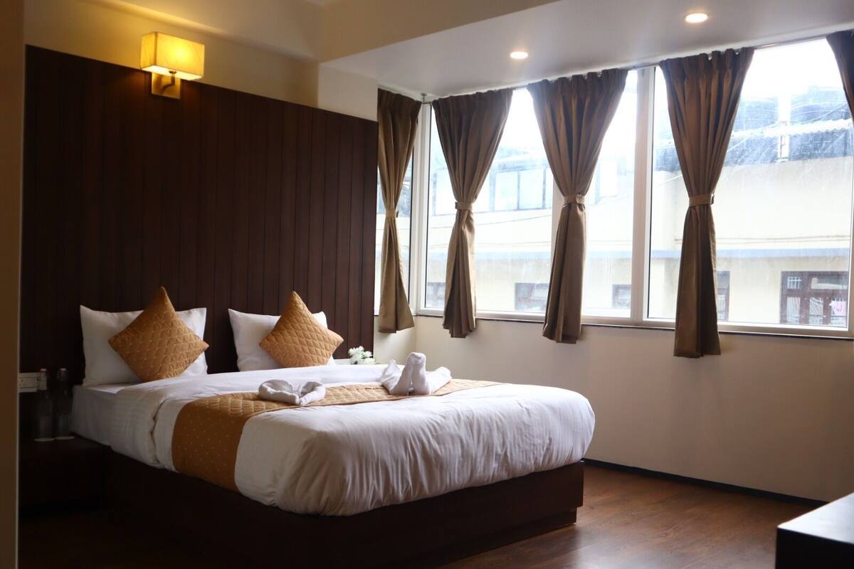 Quarto de hotel com uma cama grande, cabeceira de madeira e vista da cidade através de janelas amplas com cortinas.
