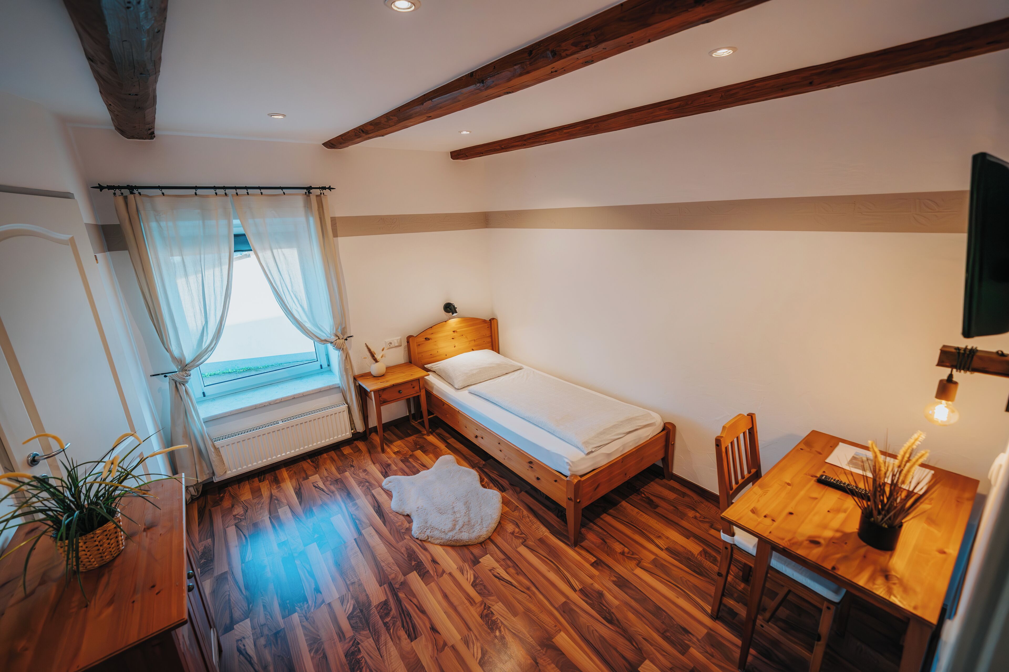 A bedroom with a wooden bed, a small table, a chair, a window with curtains, and a plant.