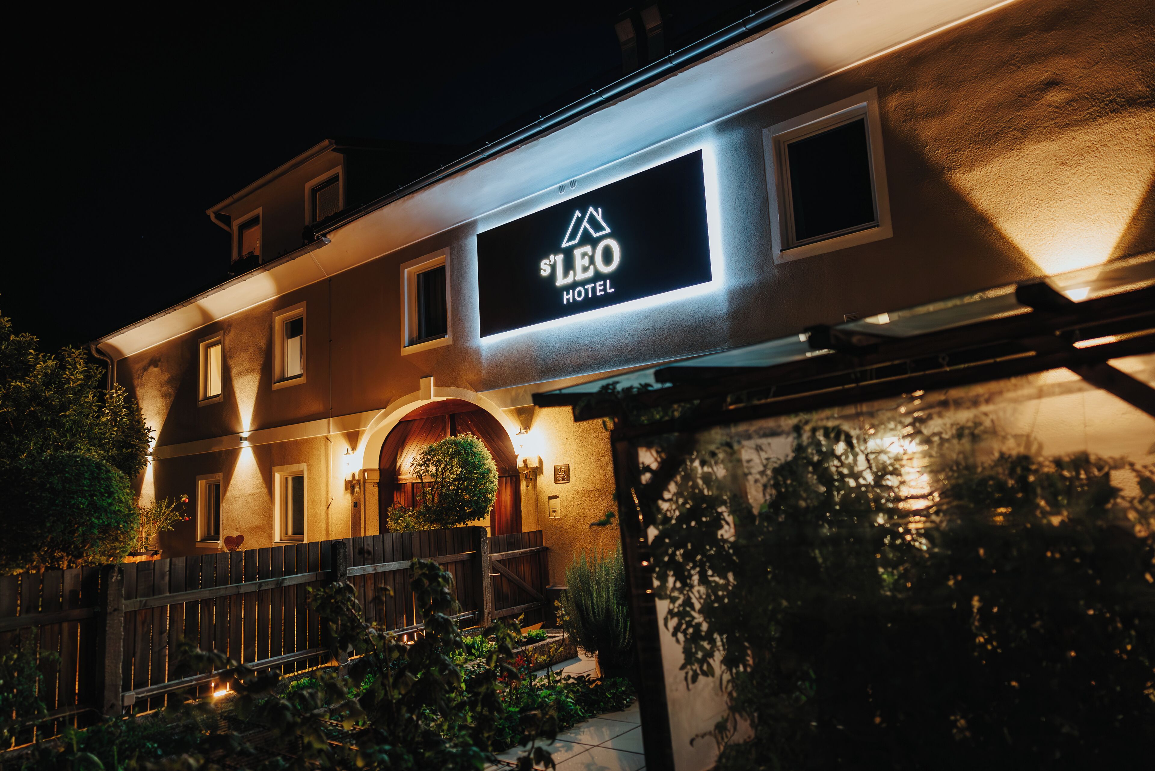 Exterior de un hotel por la noche con señalización iluminada, cerca viva de madera y un jardín.