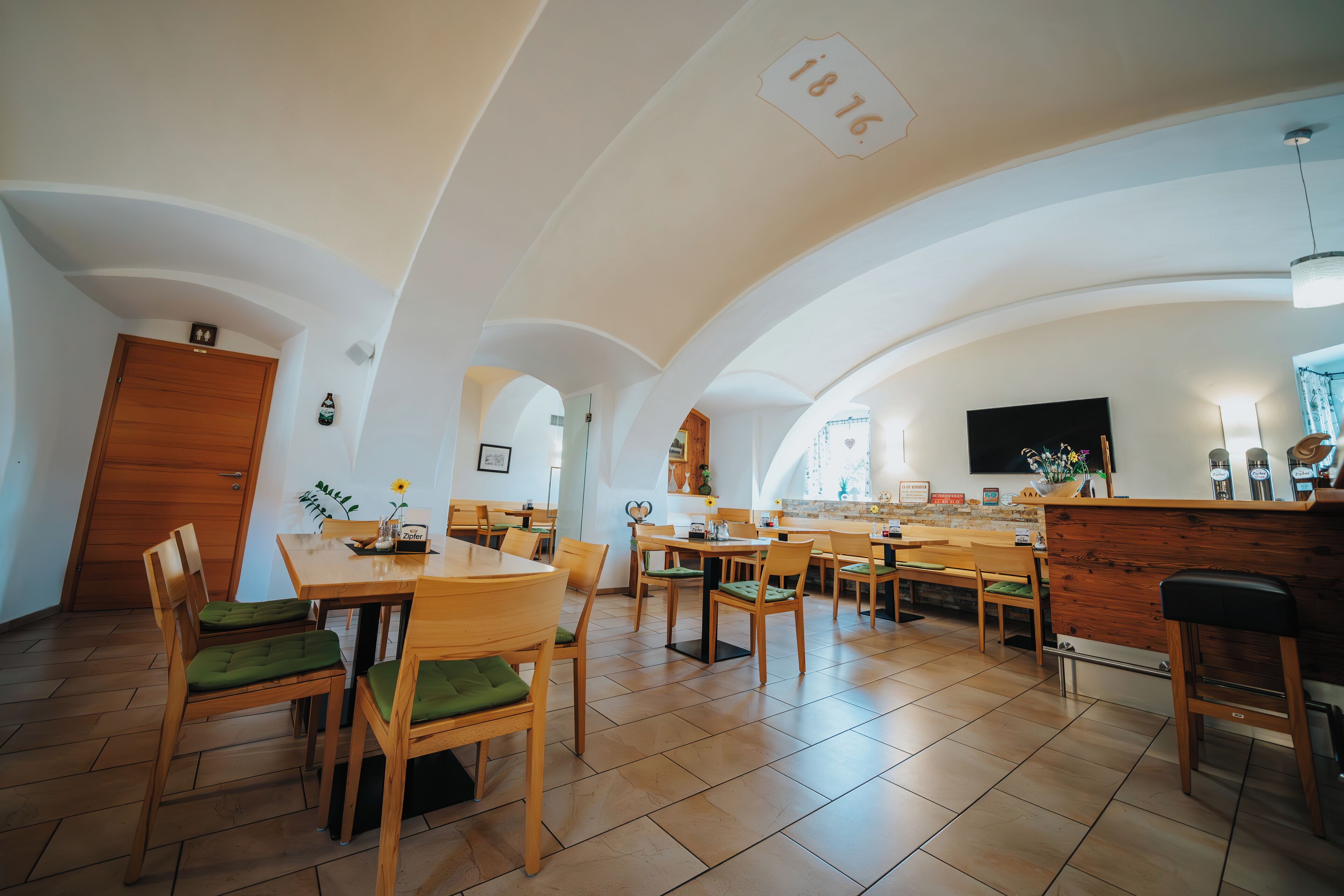 A spacious dining area with wooden tables and chairs, a kitchen counter, and a high ceiling with exposed beams.