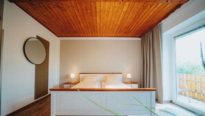 A bedroom with a wooden ceiling, a large bed with white bedding, two bedside tables with lamps, a round mirror, and a view of the outdoors through a window. - Hotel Leo (Steyr)