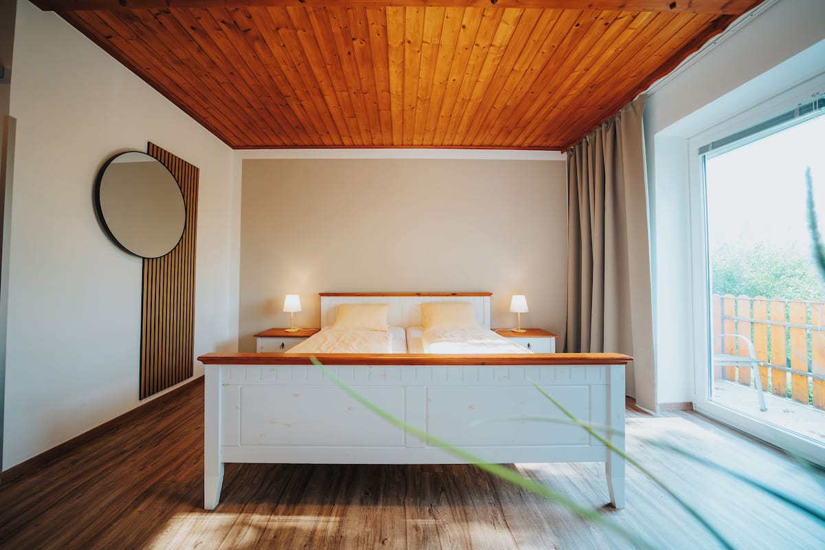 A bedroom with a wooden ceiling, a large bed with white bedding, two bedside tables with lamps, a round mirror, and a view of the outdoors through a window.