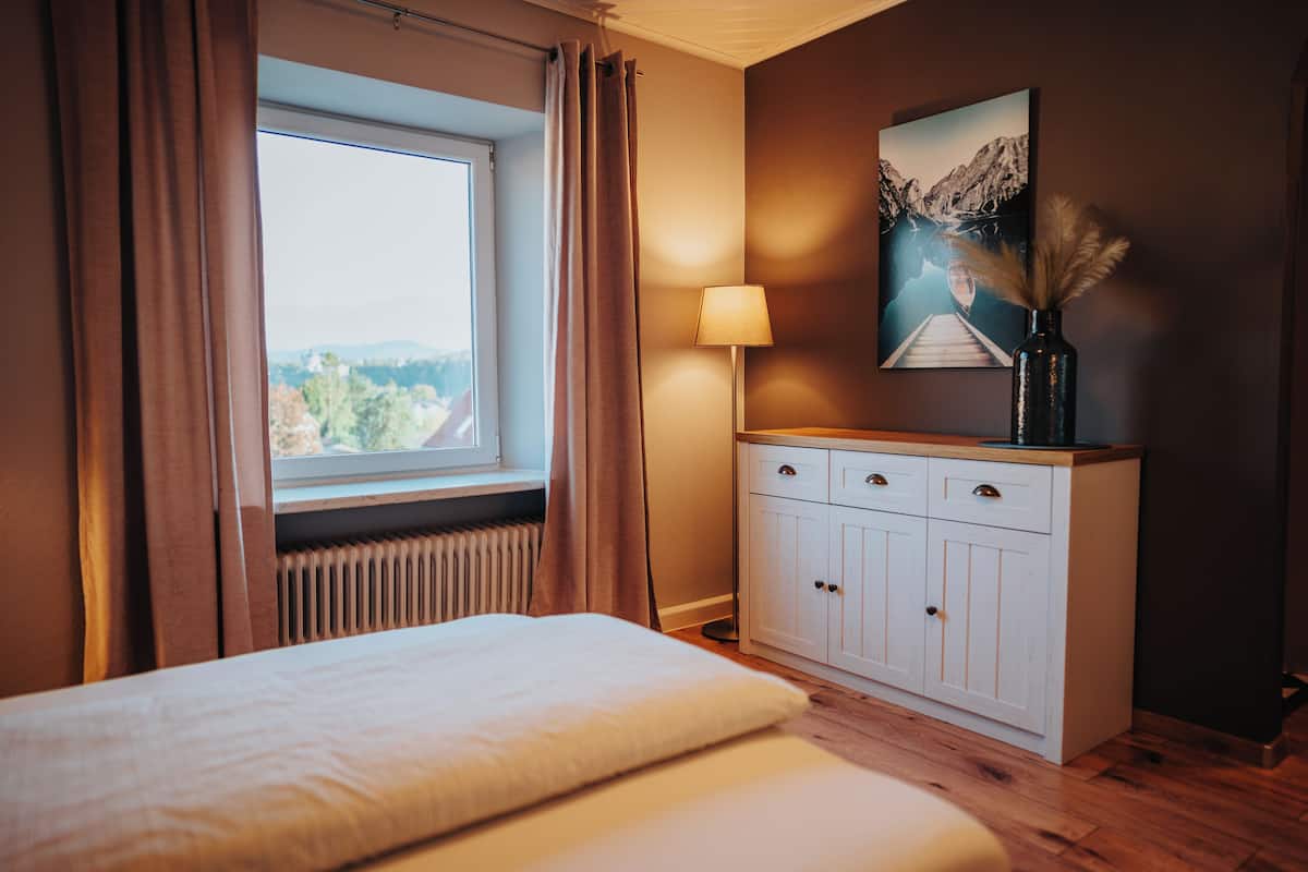 A bedroom with a bed, a window with curtains, a bedside lamp, a cabinet, and a painting on the wall.
