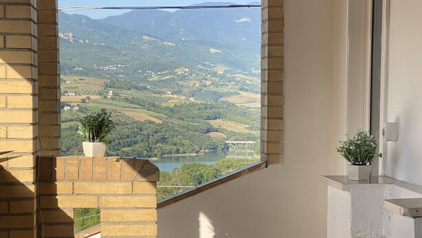 Een balkon met uitzicht op een vallei en bergen. Er staan twee planten in potten op het balkon.