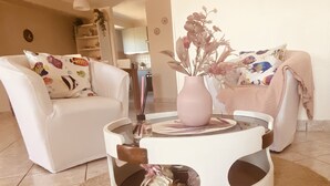 Een woonkamer met twee witte banken, een ronde salontafel met een bloemenvaas, en een keuken op de achtergrond.
