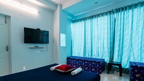 A hotel room with a bed, a TV, a window with curtains, and a small table. - Hotel Les Hues (Puducherry)