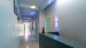 A long, narrow hallway with a reception desk on the right, a green door, and a plant on the desk. - Hotel Les Hues (Puducherry)