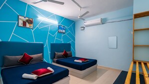 A modern bedroom with two beds, a ceiling fan, an air conditioning unit, and a wooden bench. - Hotel Les Hues (Puducherry)