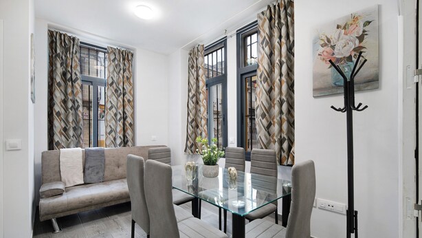 Uma área de refeições com mesa de vidro, cadeiras cinzas, um sofá e cortinas estampadas.