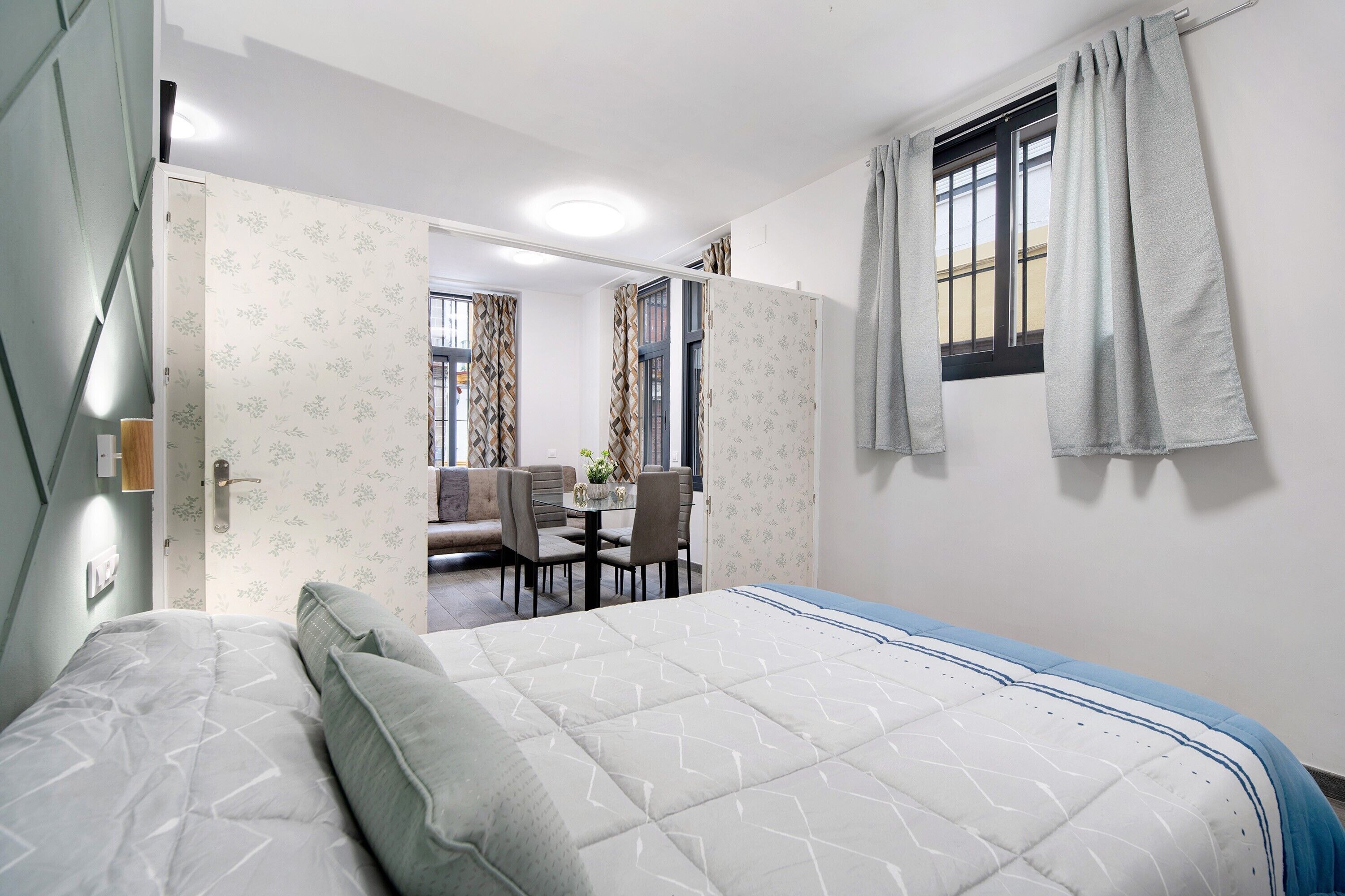 Een slaapkamer met een bed, een eettafel met stoelen, een raam met gordijnen en een deur die naar een andere kamer leidt.