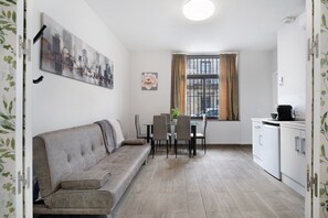 A compact living space with a sofa, dining table, chairs, a small kitchen area, and a window with curtains.