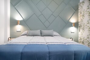 A neatly made bed with a blue and white duvet, two bedside lamps, and a patterned wallpaper on the right wall.