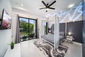 A bedroom with a canopy bed, a ceiling fan, a large window, and a view of greenery outside. - Elements Luxury Home with Pool and Hot Tub Near the Beach (Fort Lauderdale)