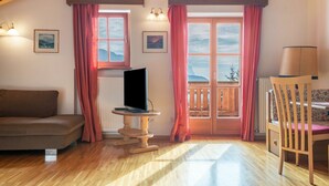 A room with a wooden floor, a brown sofa, a television on a wooden stand, a wooden chair, and a balcony with a view. - Apartment 'Funtnatsch Langkofel' with Mountain View, Shared Garden and Wi-Fi (Laion)