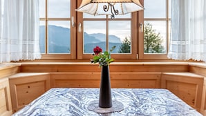 A wooden cabin room with a table, a vase with a rose, and a view of mountains through large windows.