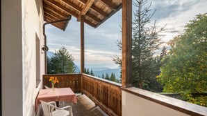A balcony with a table and chairs, a wooden railing, and a view of trees and mountains.