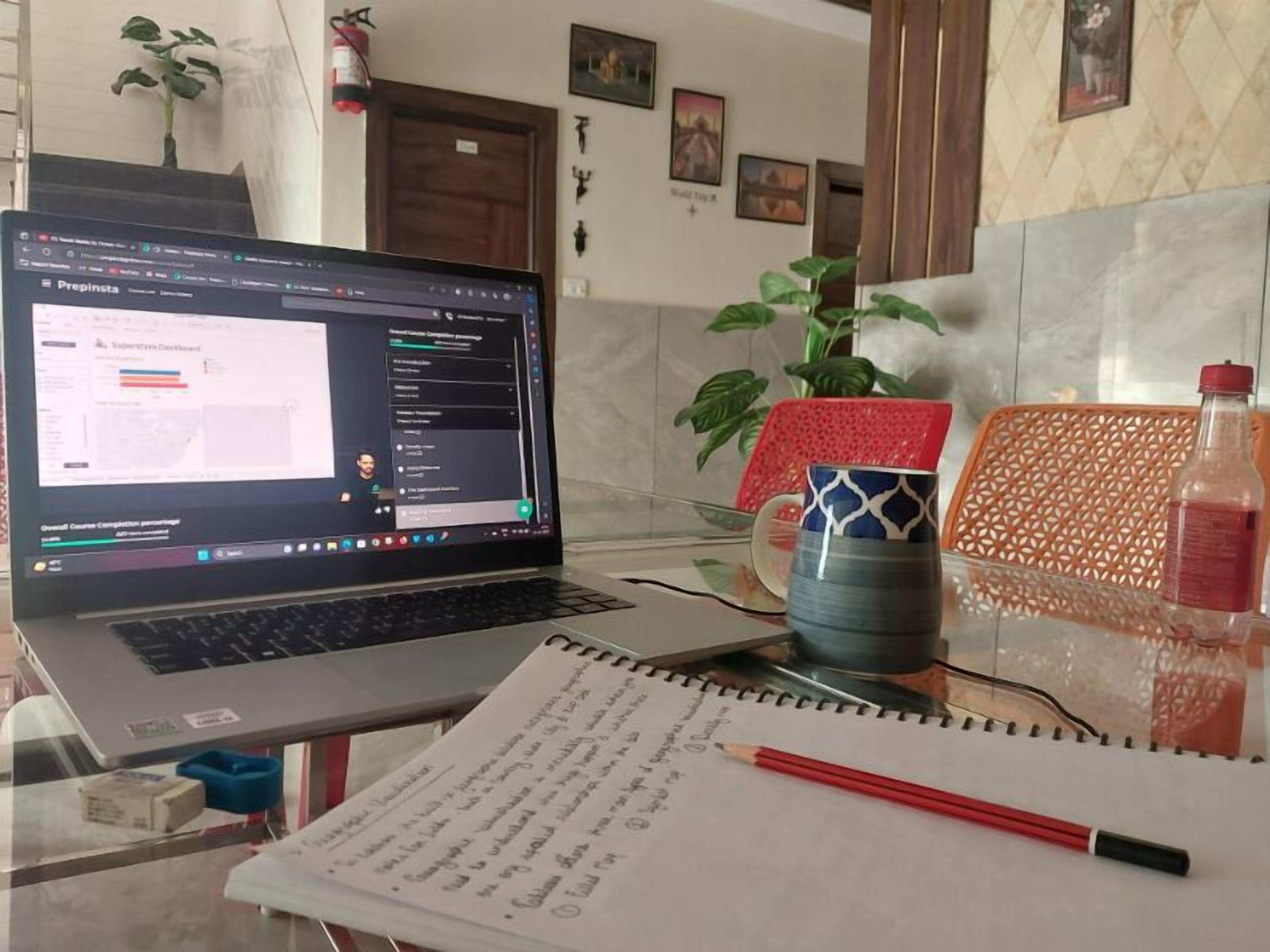 A laptop on a desk with a notebook and pencil, a mug, and a bottle.