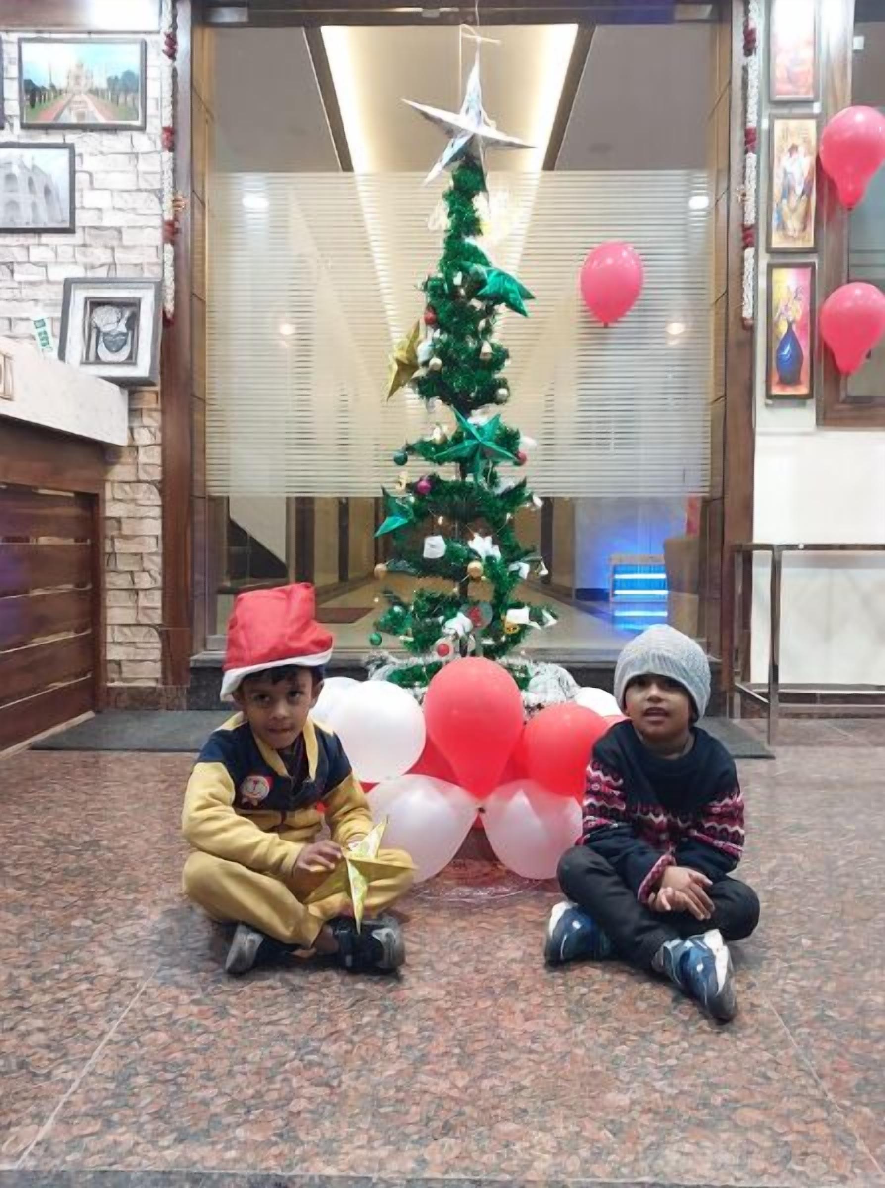 Two children sitting in front of a decorated Christmas tree with balloons.